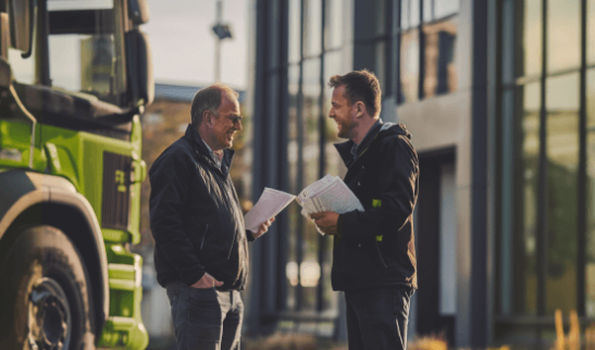 Two individuals standing by a truck, discussing over papers outside a building.